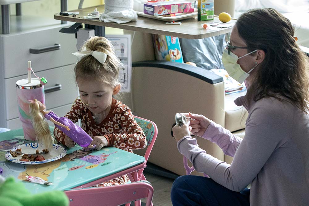 A girl with her hair in a bun plays with a princess set. She has medical tubes in her nose. A woman wearing a surgical mask sits beside her.