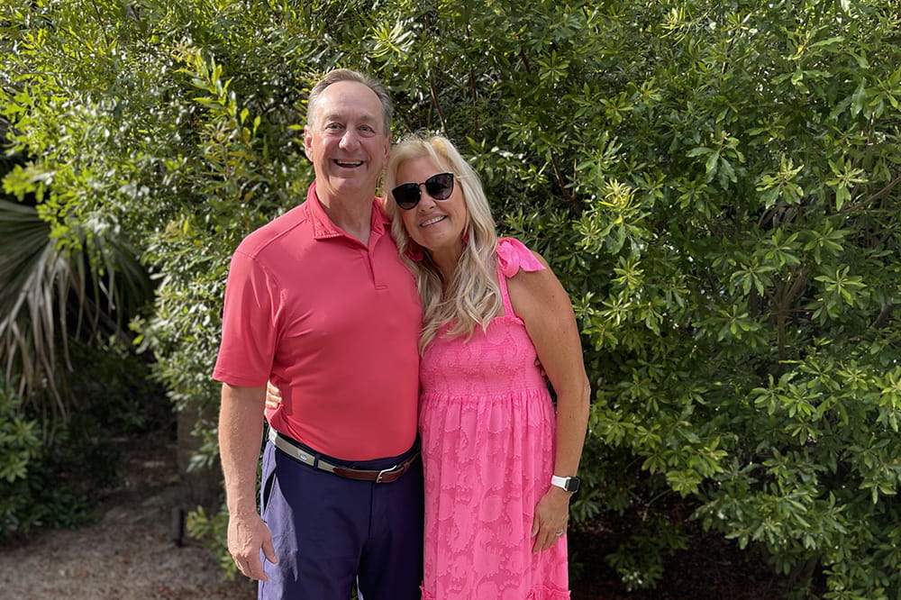 a couple,  she in a pink dress and he in a pink polo, pose in front of large green bushes