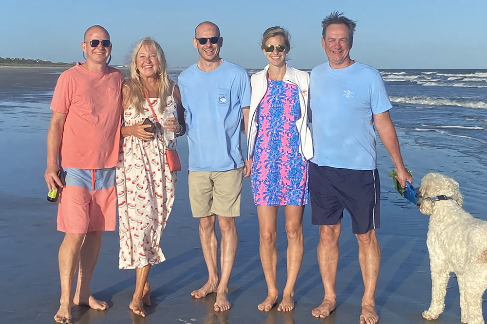 a family group of mom and dad with three adult children and a dog pose barefoot on the beach