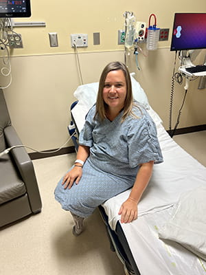 a woman in a hospital gown sits on the edge of a hospital bed looking up at the camera and smiling