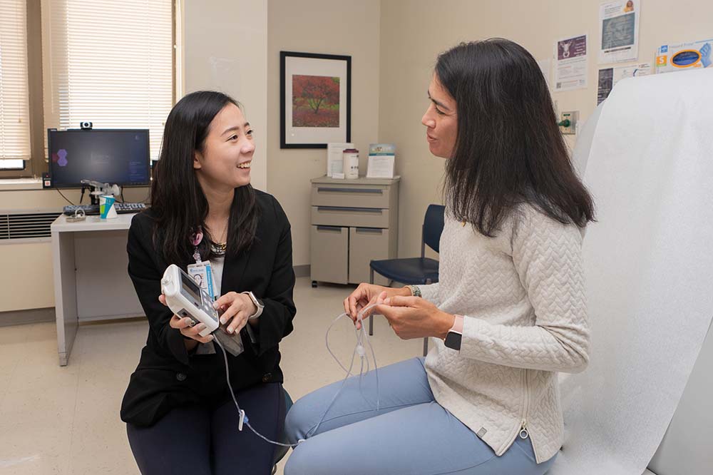 Young woman, seated to left, holds a model up to a woman who is sitting on a clinical chair.