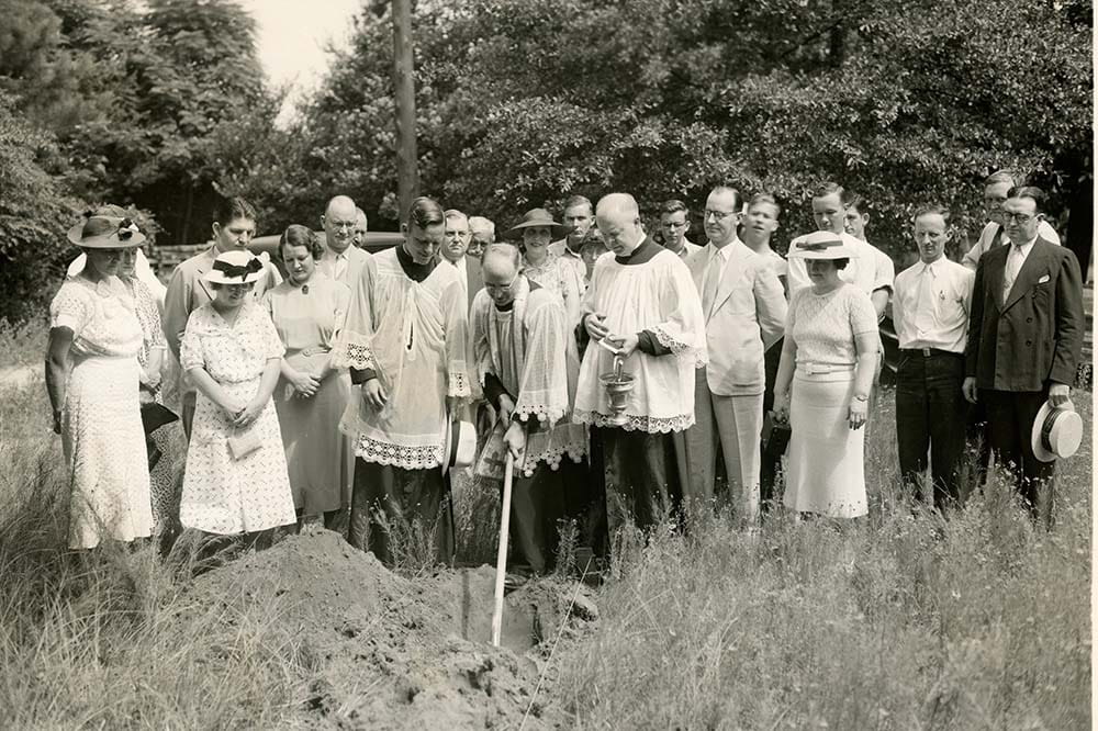 black and white photo of group of people standing outside. A man holds a shovel.