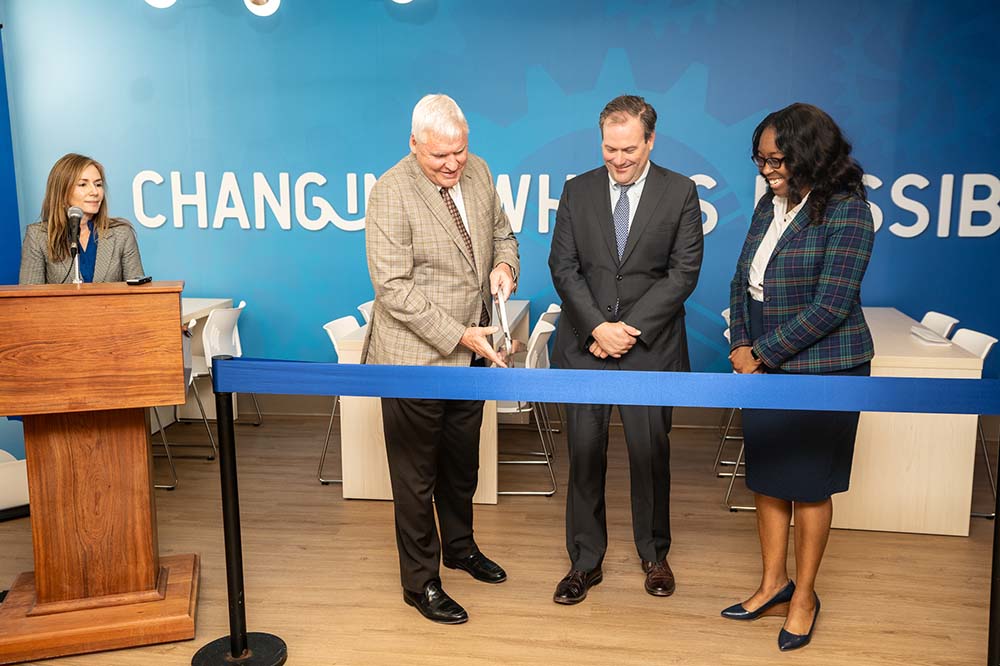 Three people stand behind a blue ribbon with a huge pair of scissors. A woman stands behind a podium to the left of the trio.