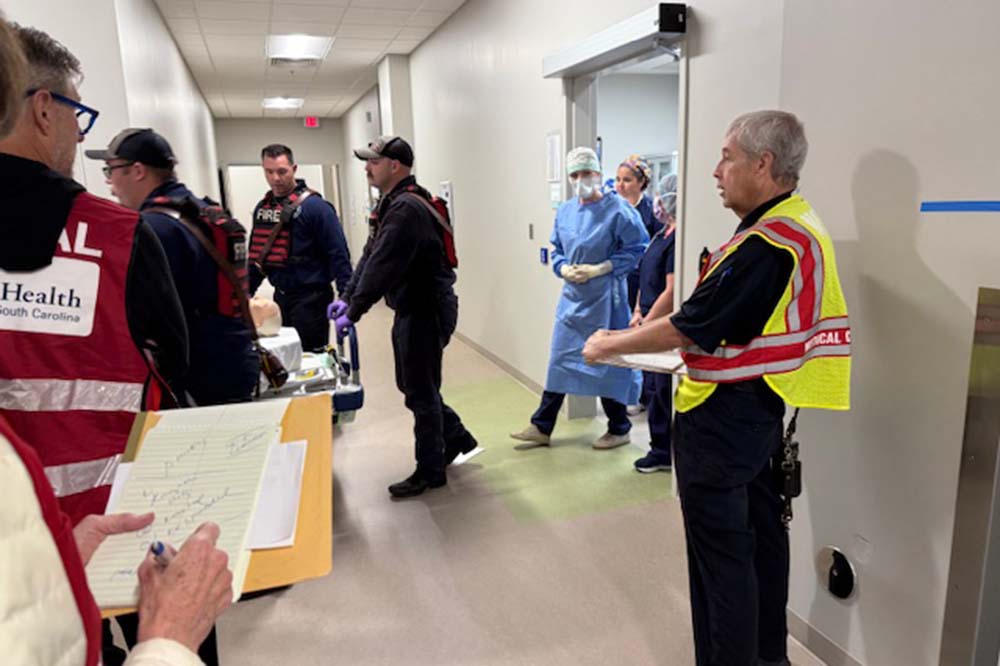 A man wearing a yellow vest looks to the left side of the picture. .There are other people gathered in a hallway, including a person in blue surgical scrubs.