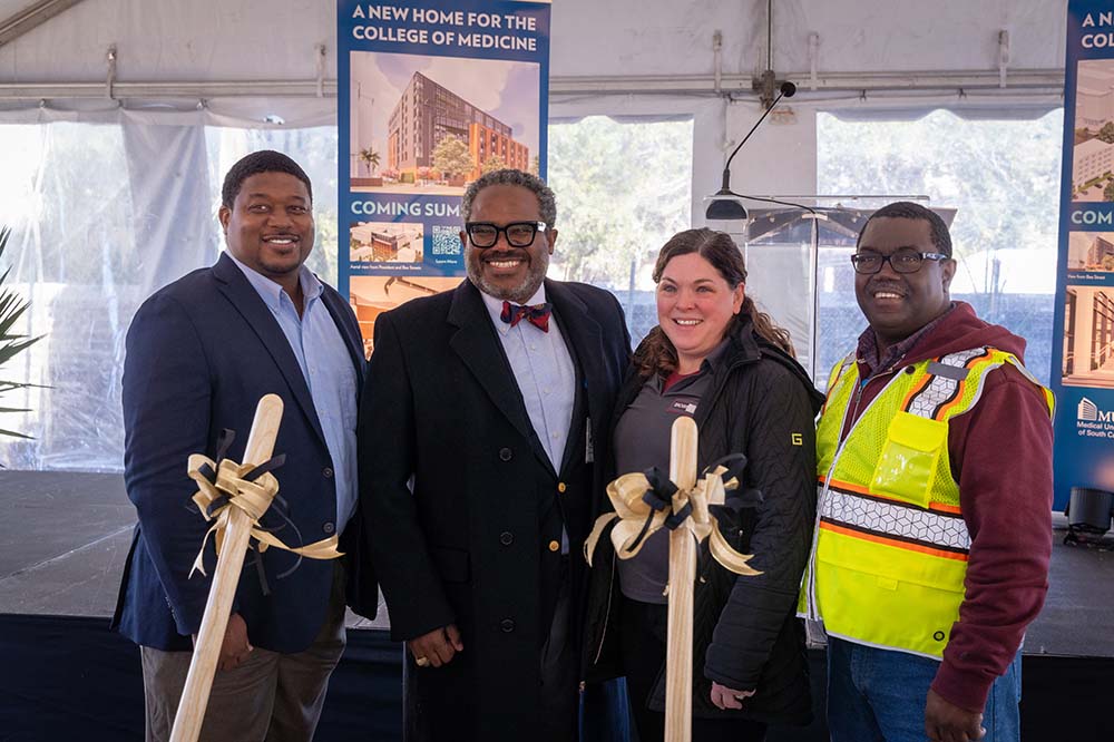 Four people smile behind two wooden shovels that have ribbons on them. They are in a white tent.