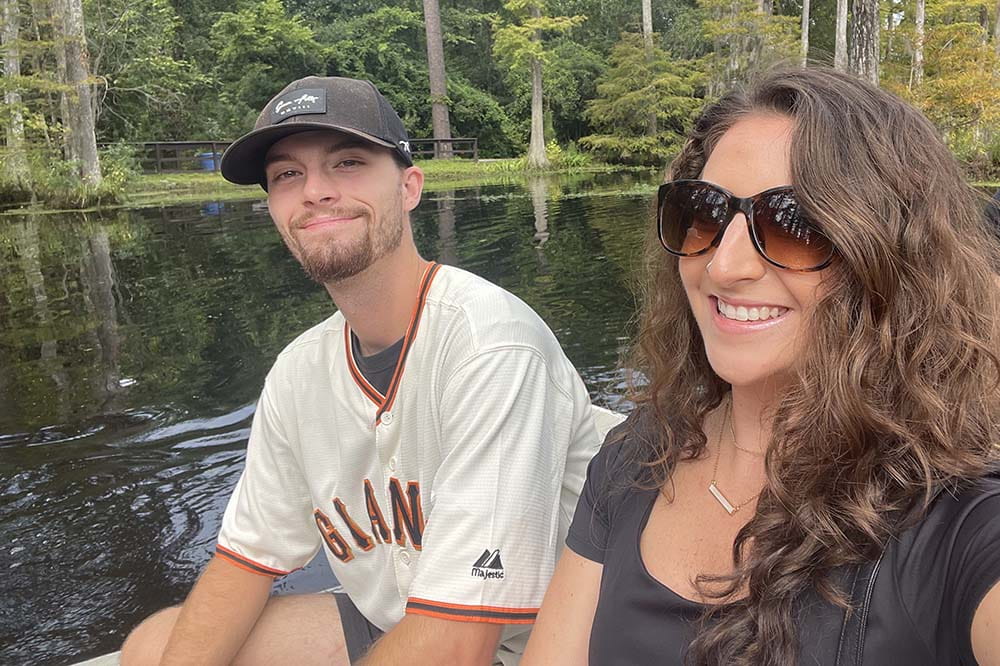 A man wearing a baseball hat and a Giants shirt sits with a woman who has long, dark, curly hair. She's wearing sunglasses and a dark colored top.