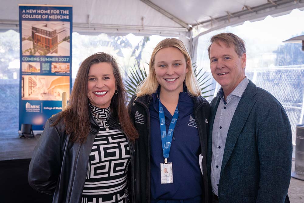 A young woman with blonde hair wearing medical scrubs is flanked by a woman and a man. They are wearing dress clothes and standing under a white tent,