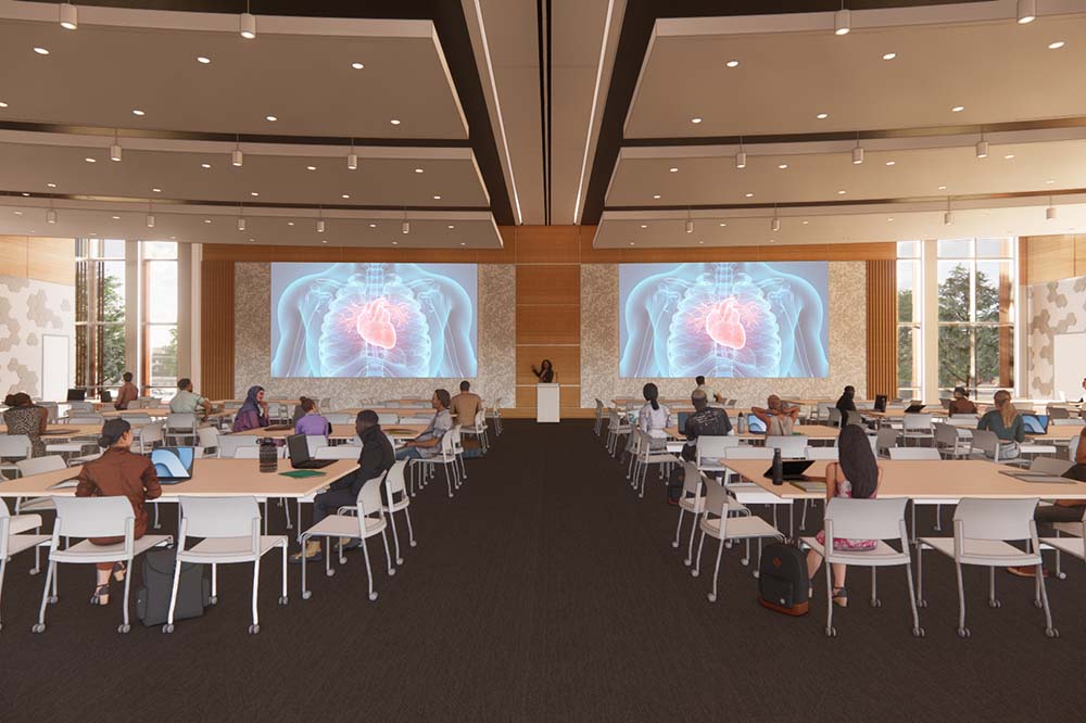 An image of a classroom with students at well spaced desks. Two monitors in front show a human torso.