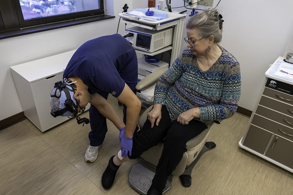 a doctor in dark blue scrubs leans down to roll up a patient's pants and look at the side of her lower leg while she sits in a chair