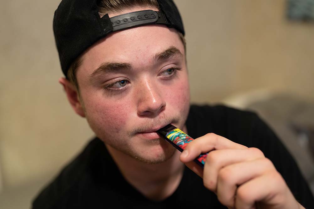 Young person vaping. The paper is wearing a baseball hat backward and a dark shirt.