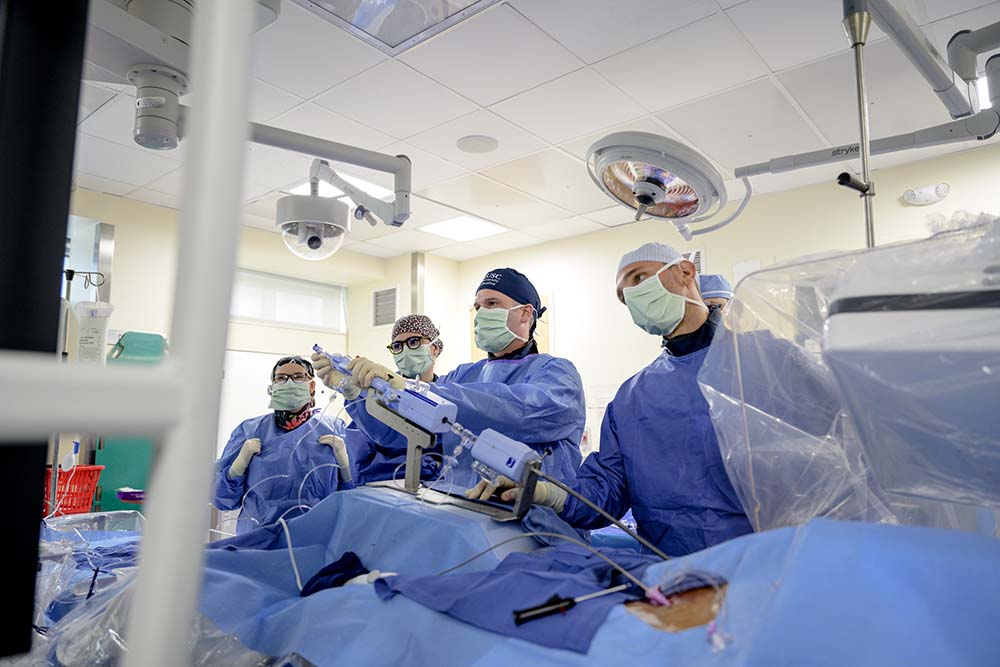 four men in blue surgical scrubs and masks lean over a blue covered table.