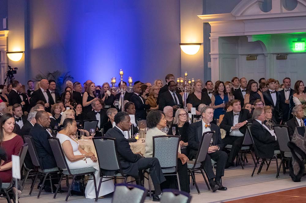 People seated and standing listening to someone out of frame. They are in a ballroom.