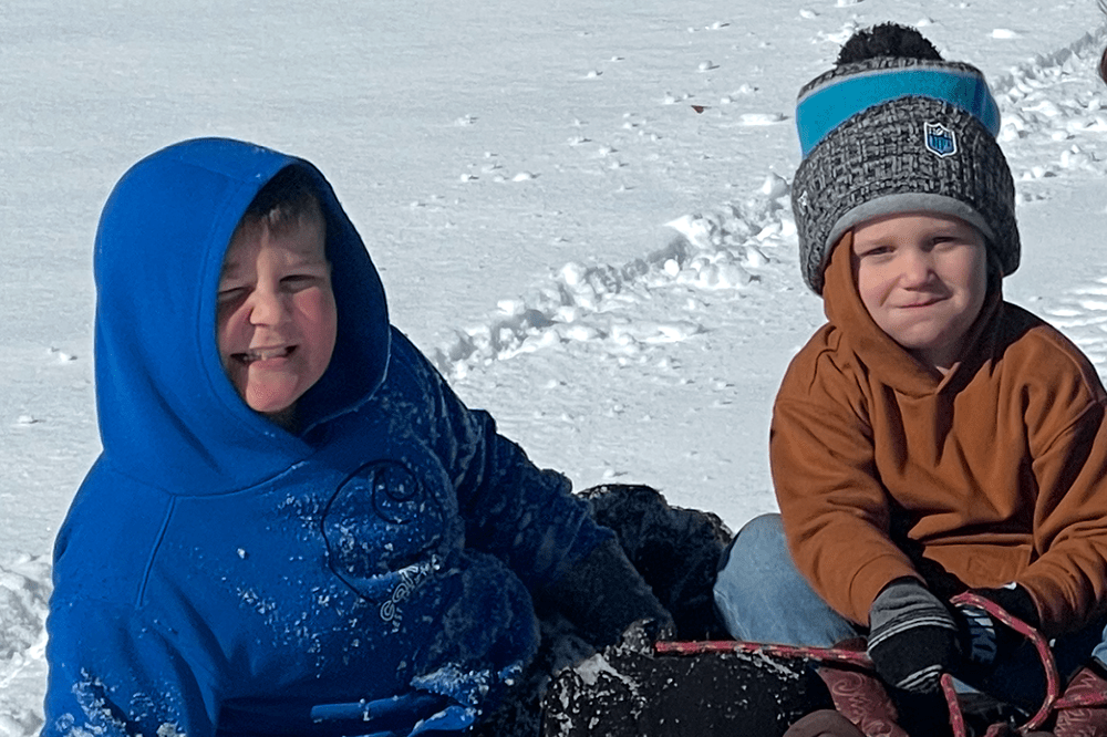 two young boys squint into the camera as they sit on a sled in the snow