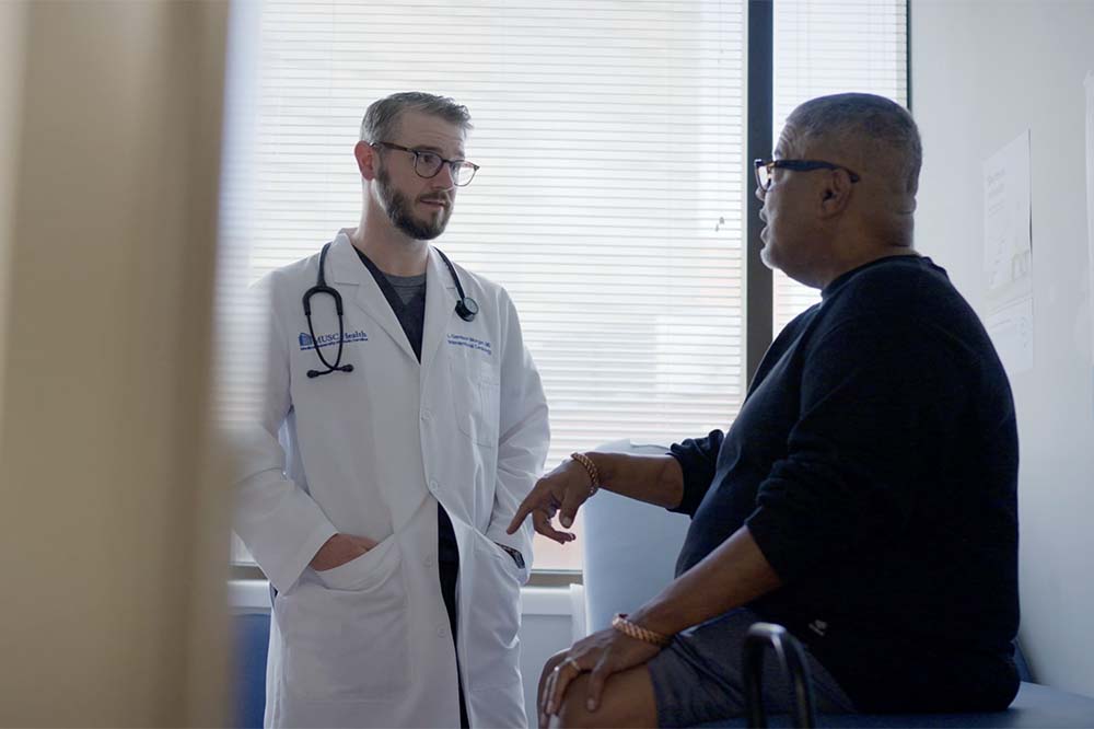 A man in a white doctor's coat with a stethoscope around his shoulders speaks to a man who is casually dressed and seated.