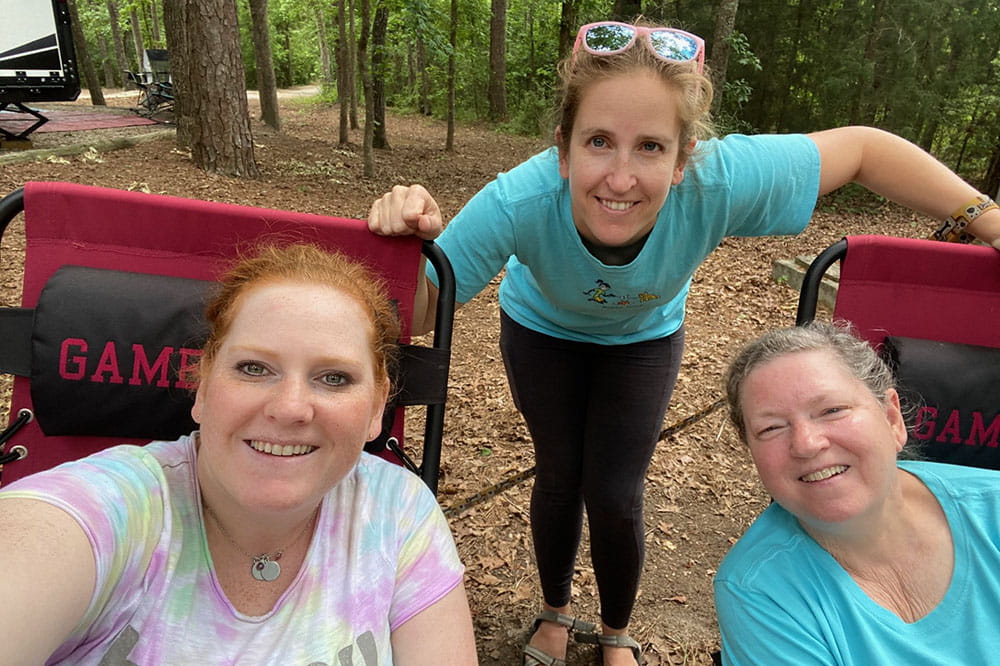 three women in casual clothing in a woodsy setting pose for a selfie