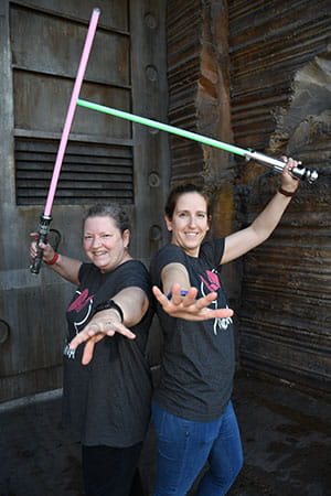 two women with huge grins pose with light sabers pulled back above their heads ready to strike at imaginary foes