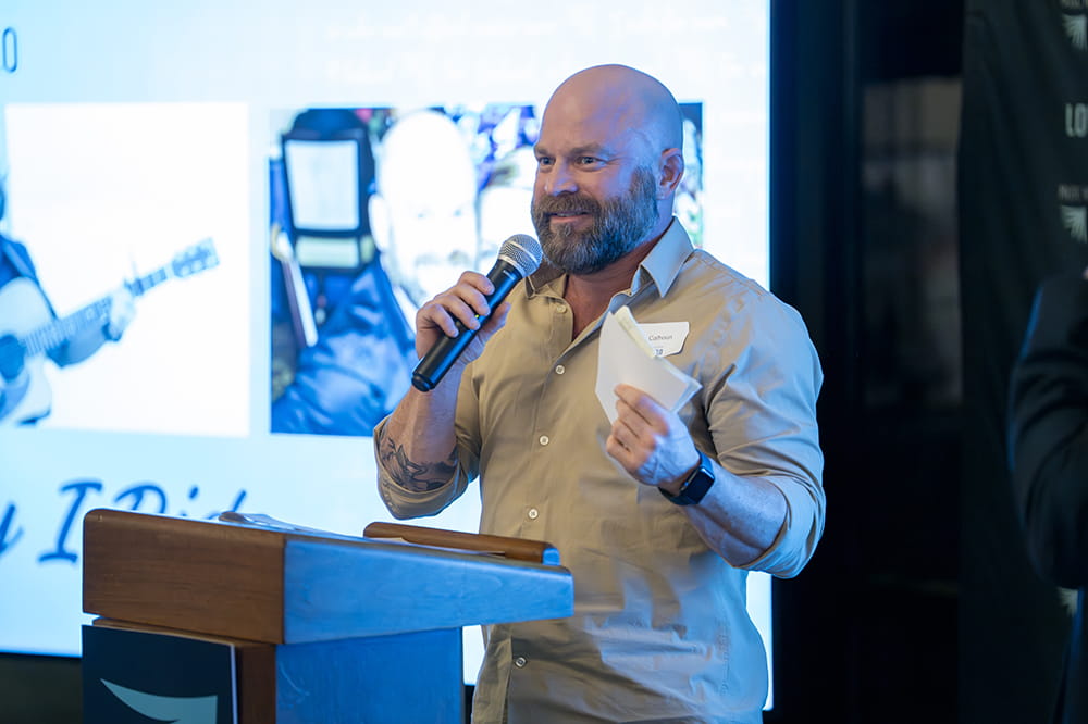 a bald man with beard stands at the podium with a microphone and a huge smile