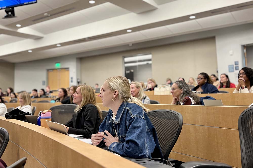 People seated at desks look to the left at an unseen speaker.