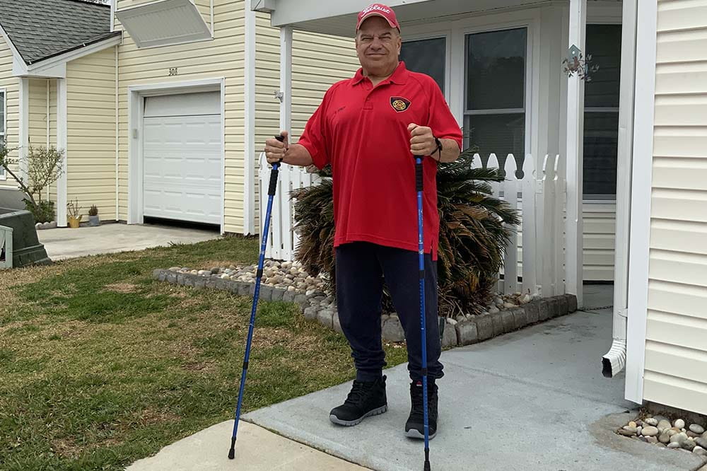A man wearing a red shirt and ball cap stands outside of a house. He is holding two walking sticks.