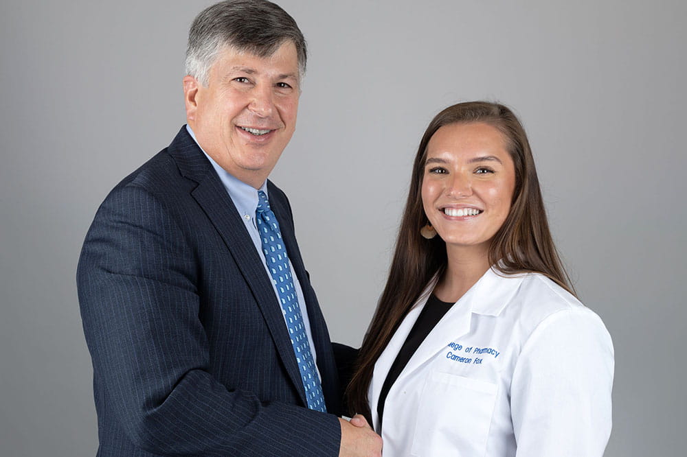 A man wearing a dark suit with a blue tie shakes hands with a young woman wearing a white coat. Both are smiling.