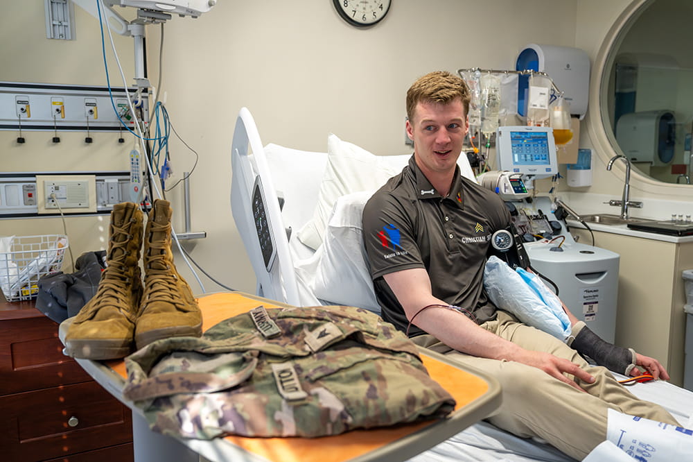 a young man in an Army green polo sits in a hospital bed with an IV in his arm with his combat uniform and boots resting on the table in front of him