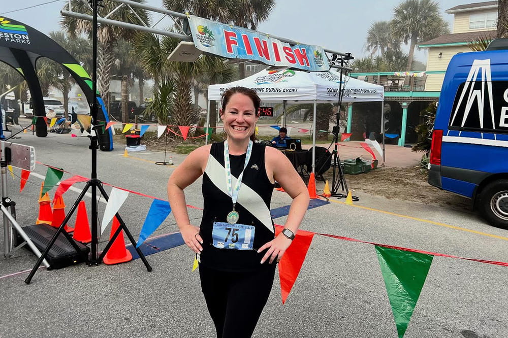 a woman in running gear poses in front of a finish line with a medal around her neck