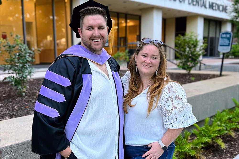 A man in graduation garb, including a cap and robe with purple accents, stands with a woman in front of a building that says James B. Edwards College of Dental Medicine.