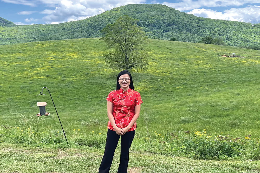 Woman wearing red top and black pants stands in front of rolling green hills.