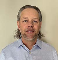 Closeup of man with longish hair and a goatee. He is wearing a blue collared shirt.