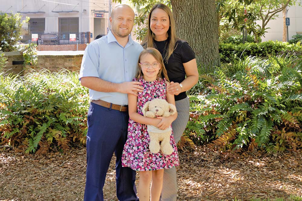 A man, woman and girl stand together in front of a tree. The girl is wearing a dress and holding a stuffed animal.