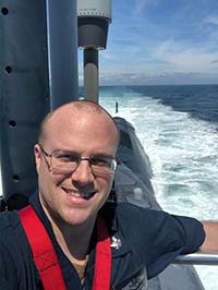 Man smiles while standing on a ship with water behind him