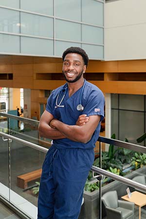 Man wearing blue hospital scrubs leans against a rail while smiling.