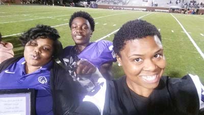 Young man on football field wearing a purple jersey with two women standing in front of him smiling.