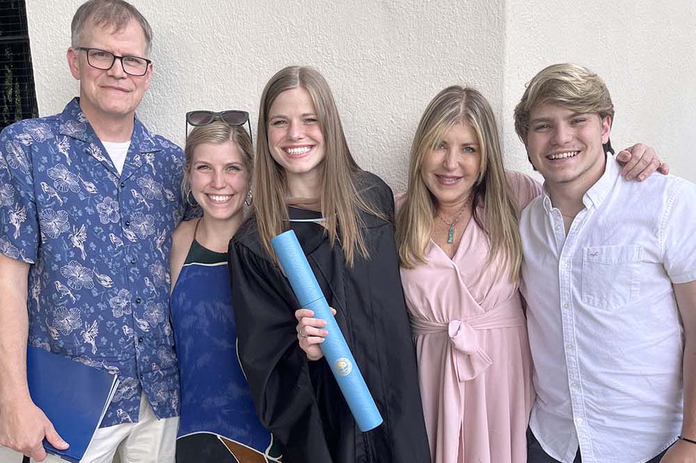 A man, three women and a younger man stand together and smile. The women in the middle is wearing a graduation robe and holding a rolled document.