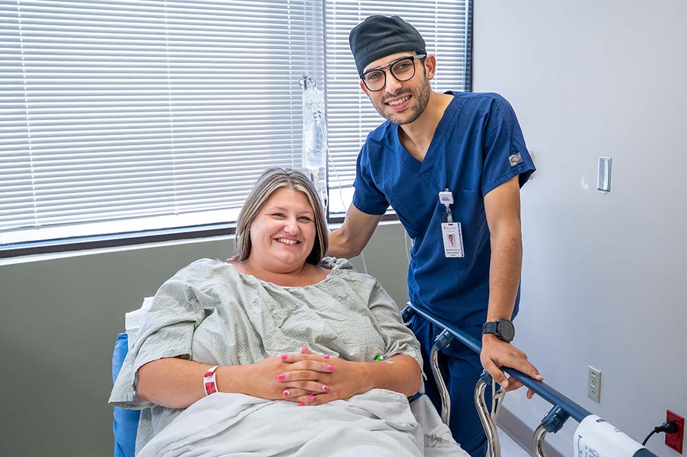 A women lying in a hospital bed with a man in blue scrubs standing beside her.