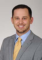 Headshot of man smiling. He's wearing a grey suit, blue shirt and yellow tie.