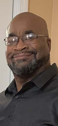 Smiling man wearing a dark collared shirt. He has a beard and is wearing glasses. 