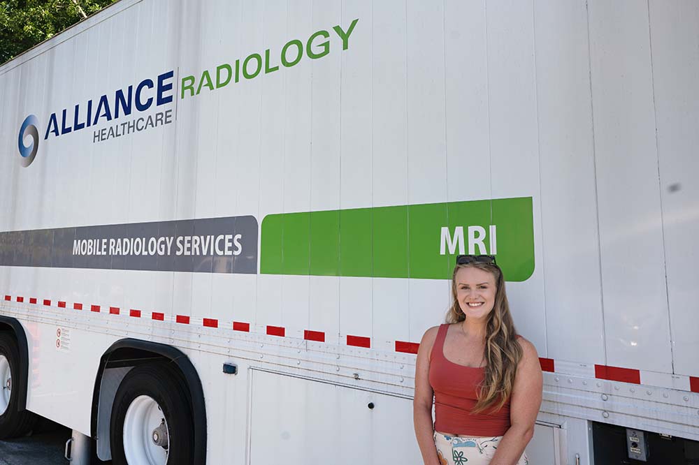 Woman with long hair and a tank top stands beside a truck that says Alliance Radiology.