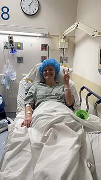 Woman lies back in a hospital bed and makes the peace sign. She has a blue cap on her head and she's under sheets.