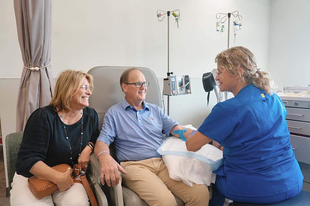 A woman and a man are seated in a clinic. He is connected by tubes to an IV pole. Another woman in blue scrubs talks with them. All three are smiling.