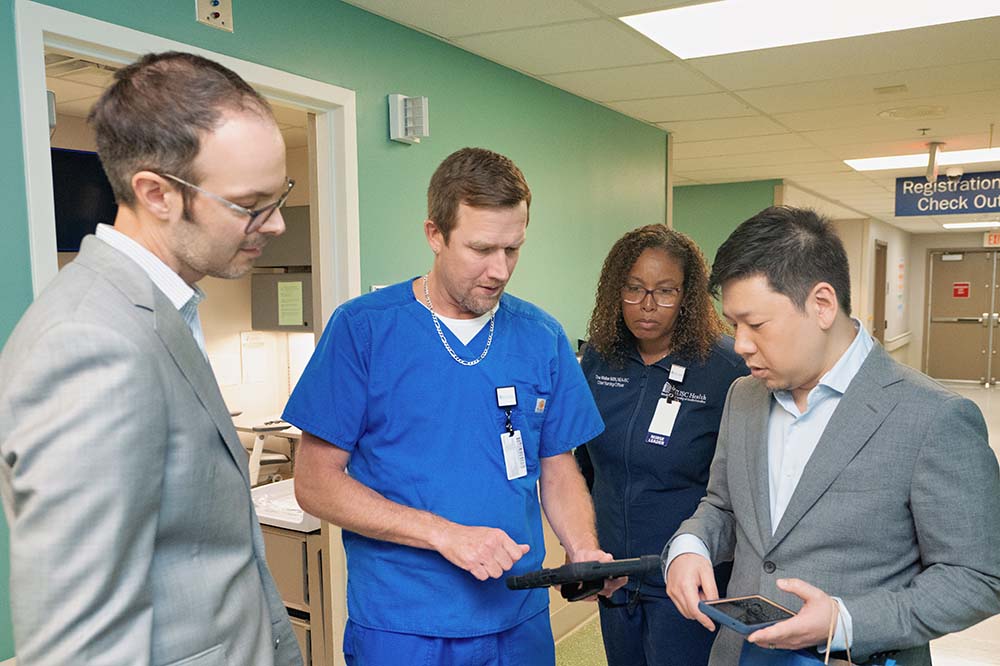 Three men and one women look at a tablet in a hallway. Two men are wearing suits. The other is wearing scrubs, as is the woman.