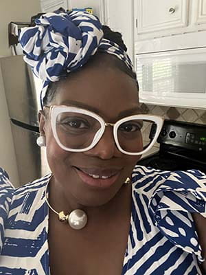 Smiling woman wearing a headscarf and matching top. She's also wearing white framed glasses.