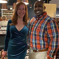A woman with red hair wearing a blue dress stands with a man wearing a plaid shirt.
