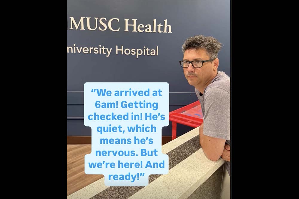 Man leans on a counter in a room with the words MUSC Health University Hospital on the wall. There are more words superimposed on the photo, saying We arrived at 6am! Getting checked in! He's quiet, which means he's nervous. But we're here! And ready!