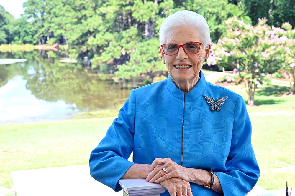 Smiling woman with white hair is wearing a blue jacket and glasses. She is in front of a body of water and trees.