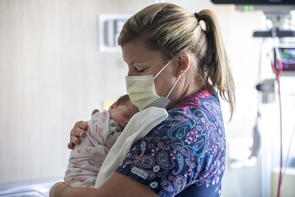 A woman wearing a surgical mask and blue scrubs holds a tiny baby to her chest.
