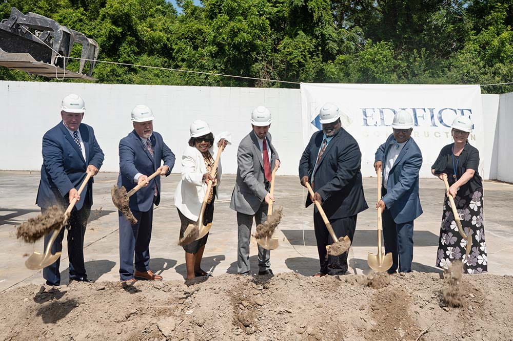Seven people wearing white hard hats lift shovels filled with dirt. A sign behind them says Edifice.