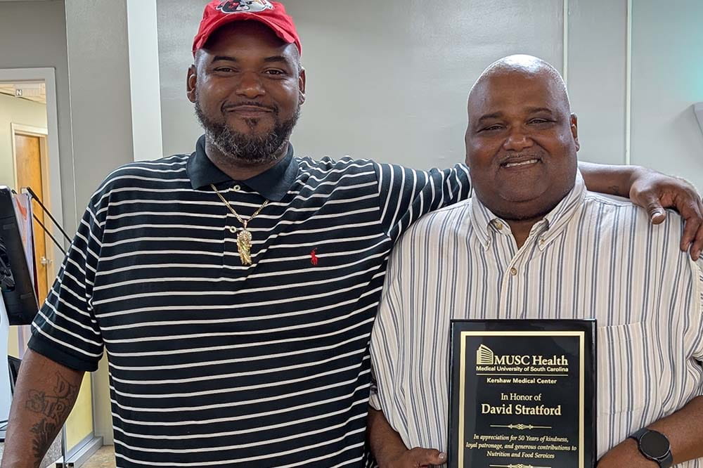 A man wearing a red ball cap and a striped shirt stands with his arm across the shoulder of another man, who is wearing a white striped dress shirt and holding a retirement plaque.