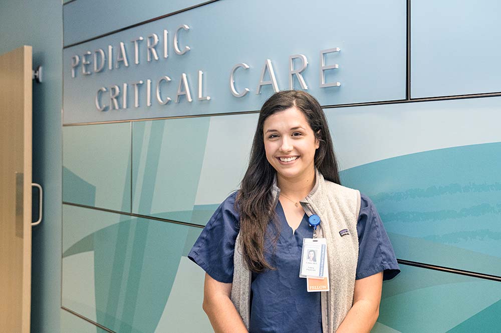 Young woman with long dark hair wearing blue scrubs smiles standing by a sign that says pediatric critical care.