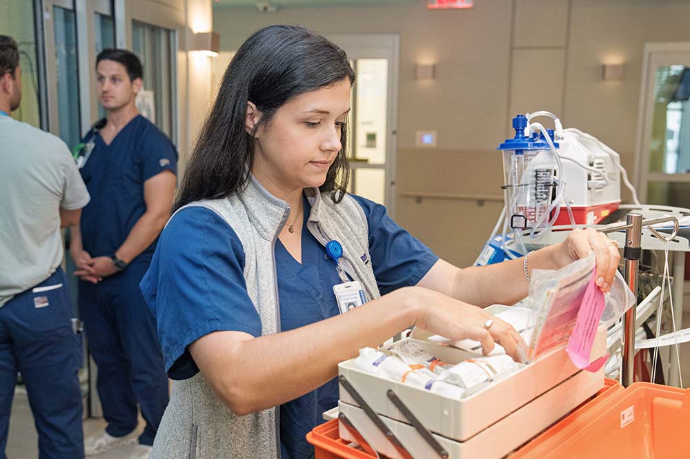 Woman with long dark hair wearing scrubs and a vest looks at a small box. There are two men in the background.